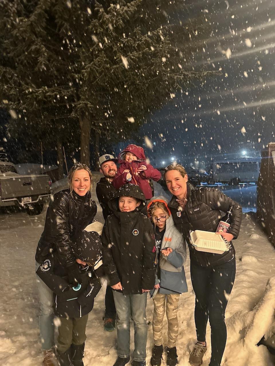 Snowy night with a group of smiling people, including children, under falling snowflakes.