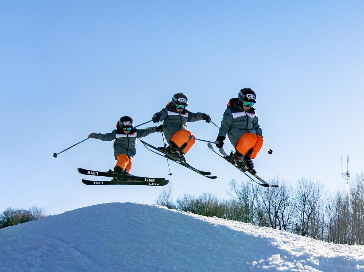 Skiers in orange pants jumping over snowy hill under blue sky.