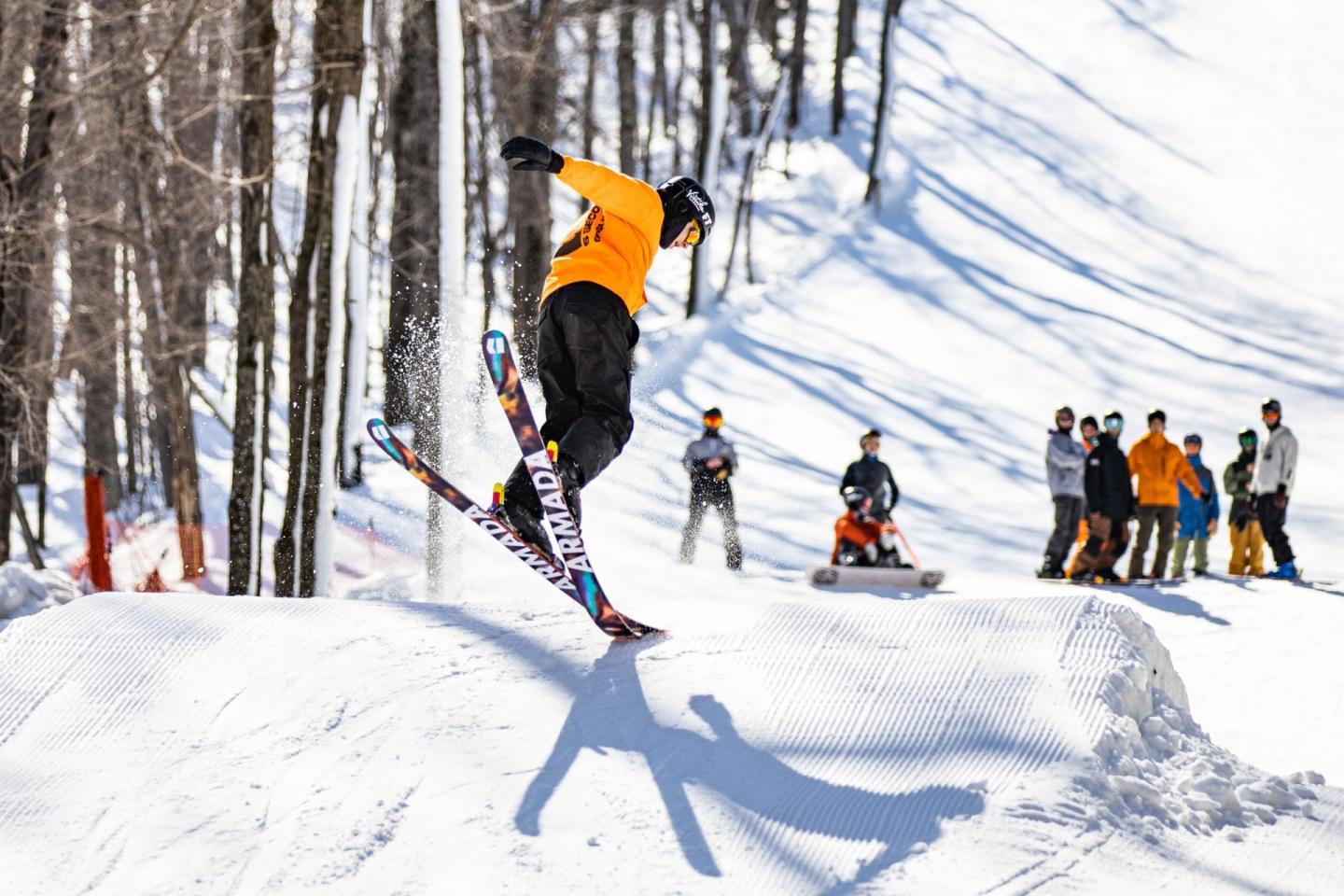Skier in mid-air jump on snowy slope, onlookers watch in background.