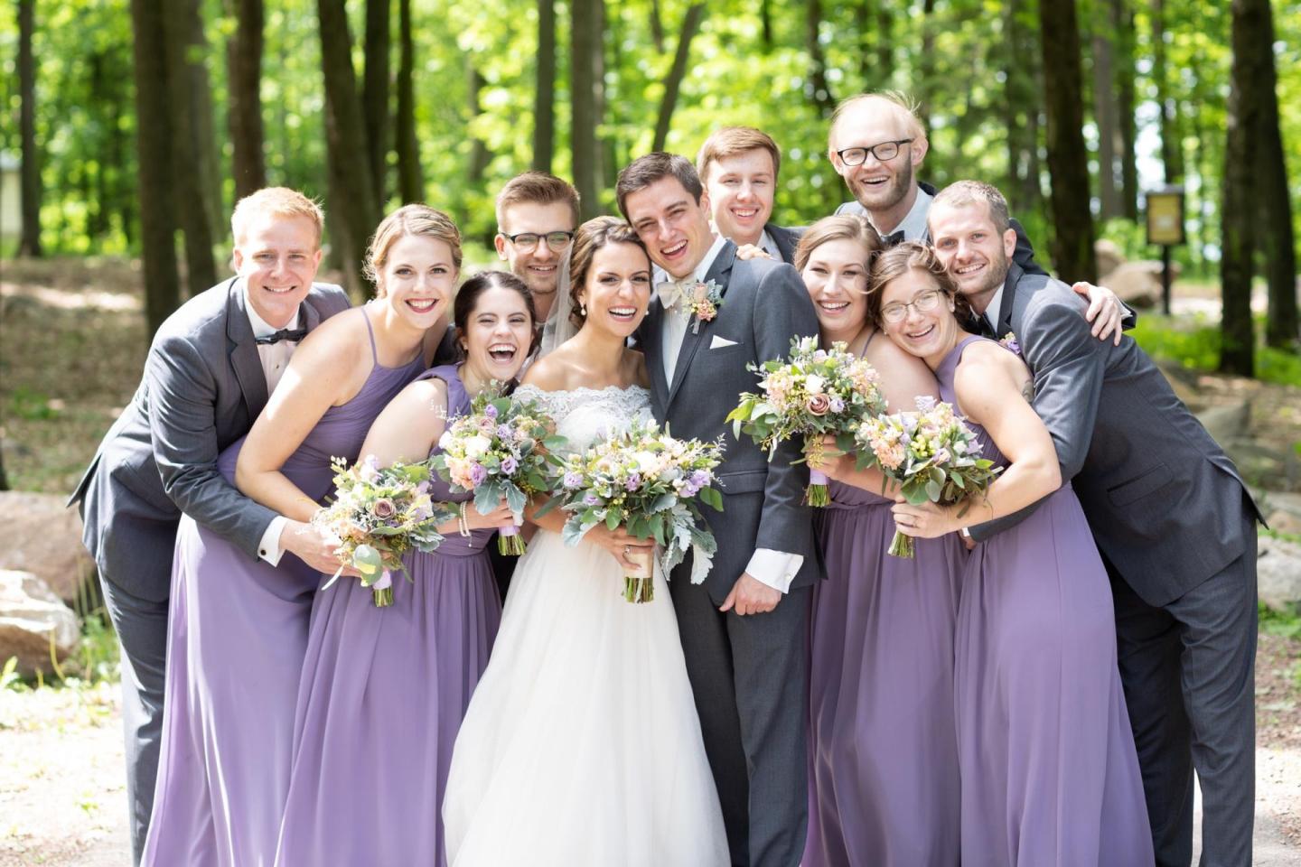 Wedding party smiling in a forest, bridesmaids in lavender, groomsmen in grey.