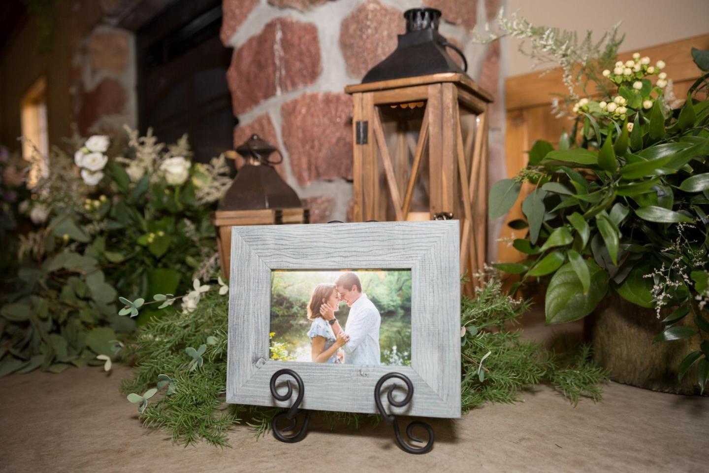 Framed photo on stand, surrounded by greenery and rustic lanterns.