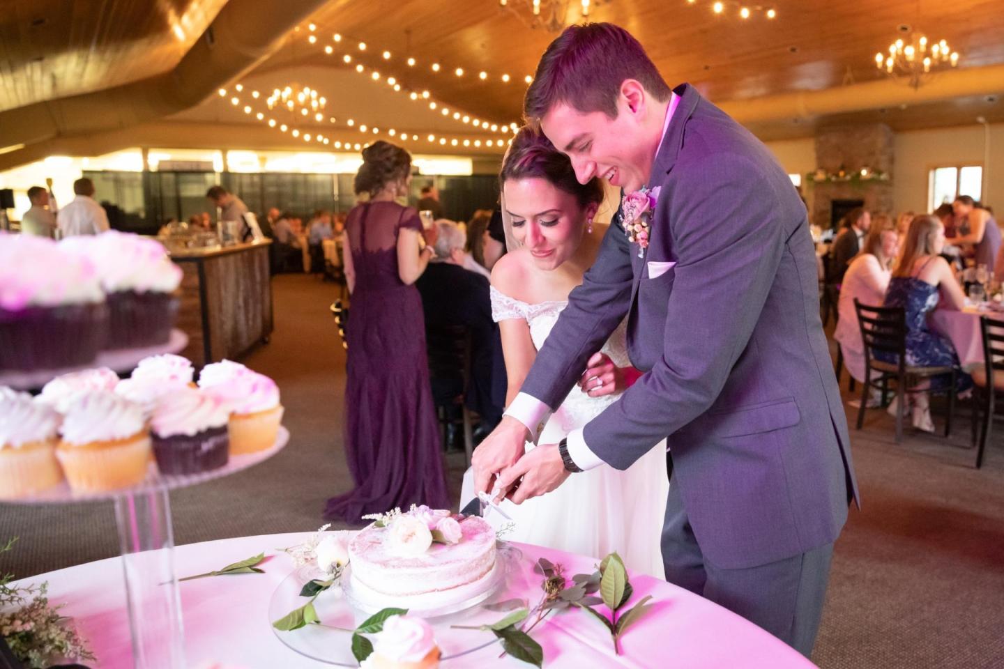 Couple cutting a cake at a wedding reception with cupcakes nearby.