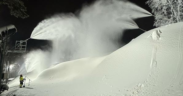 Snow machines covering a slope with snow at night; a person stands nearby.