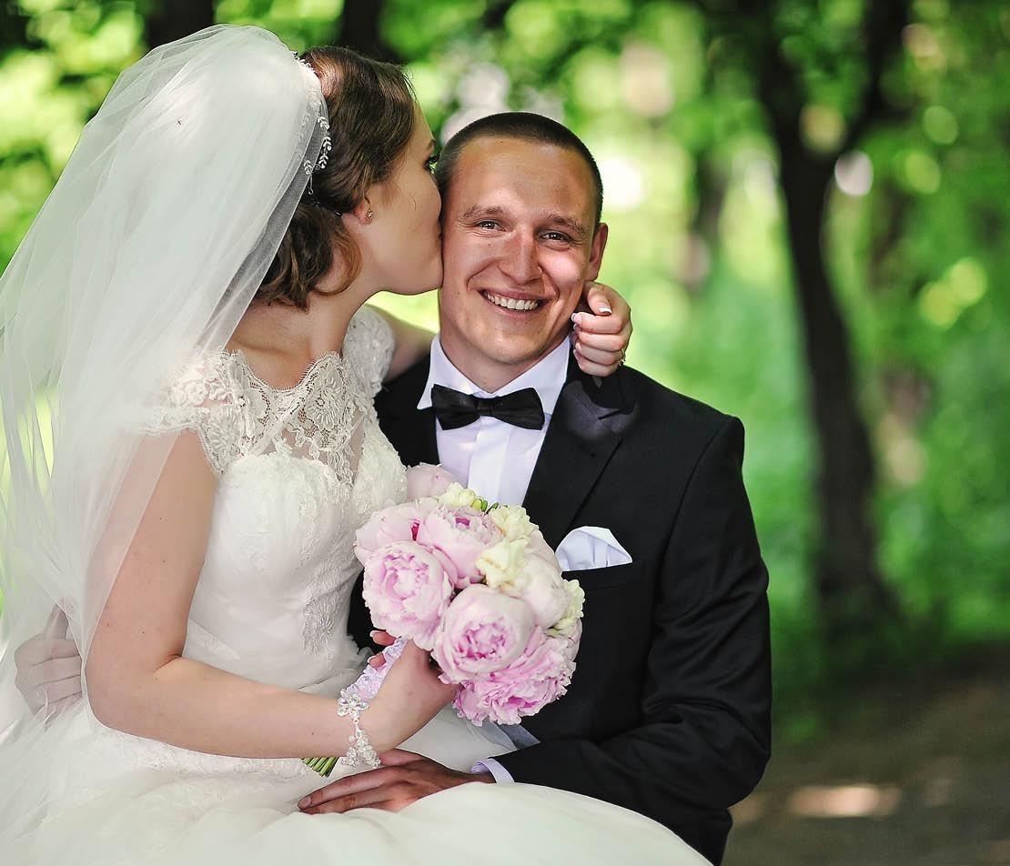 Bride kissing groom's cheek, holding pink bouquet, in a lush green setting.