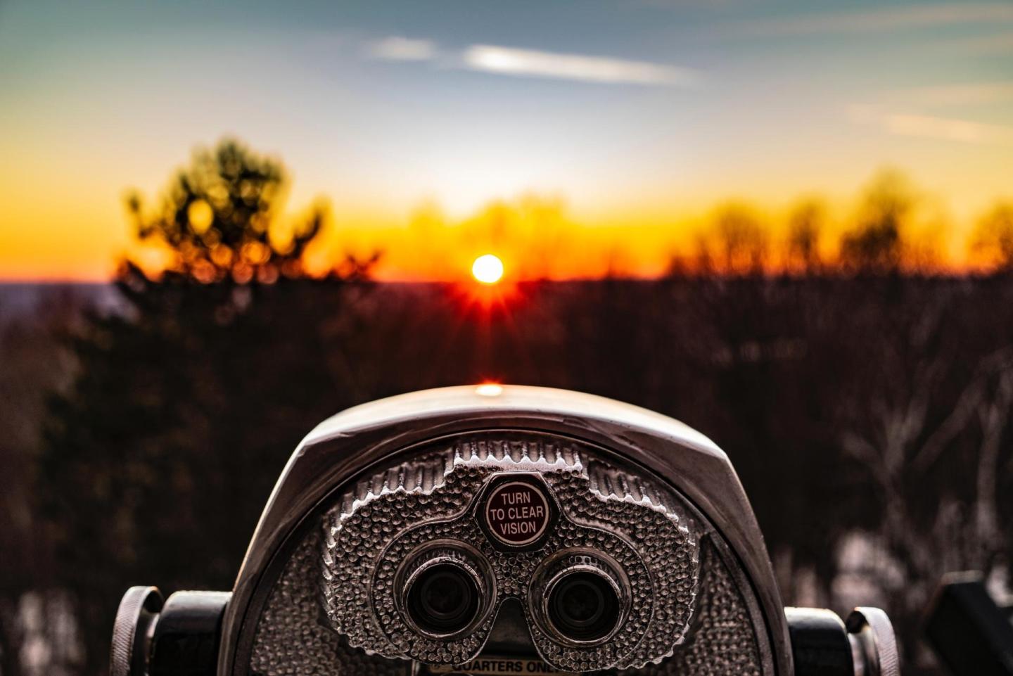 Binocular viewer facing a vibrant sunset over a forested landscape.