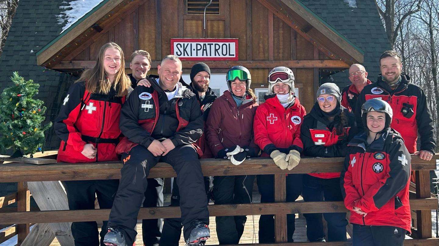 Group of skiers in red jackets smiling on a snowy wooden deck.