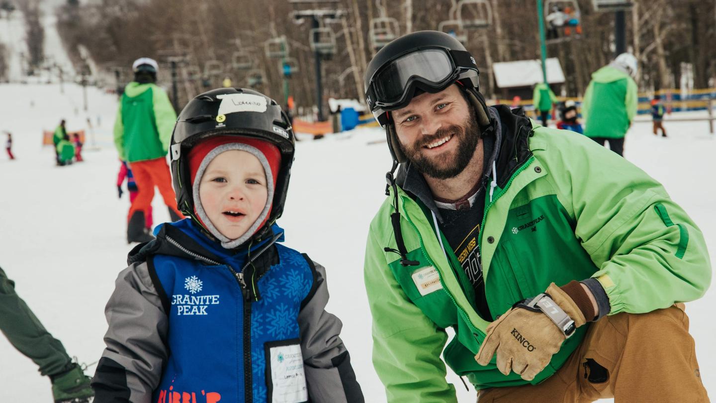 Child and adult in ski gear smiling on snowy slope.