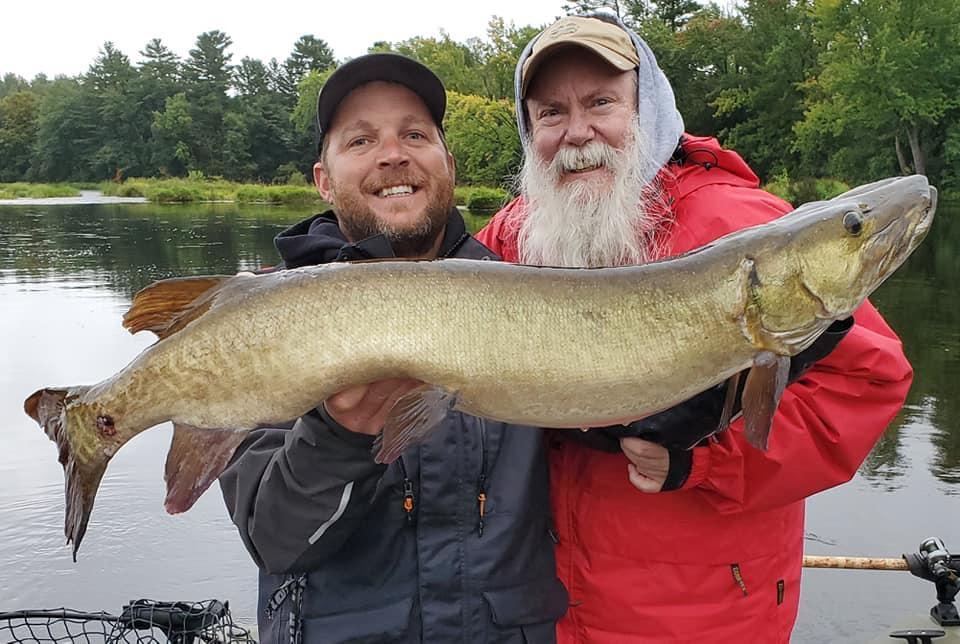 Two men proudly holding a large fish by a lake.