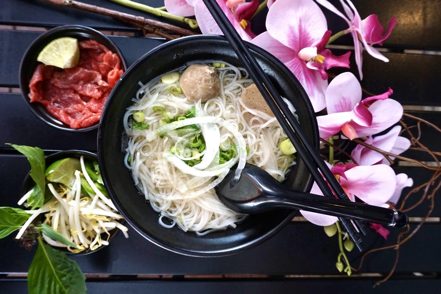 Noodle soup with meatballs and vegetables in a black bowl, garnished with pink flowers.