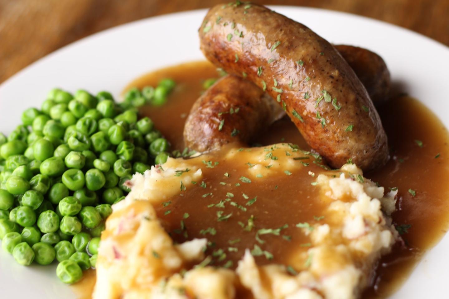 Sausages with gravy, mashed potatoes, and peas on a white plate.