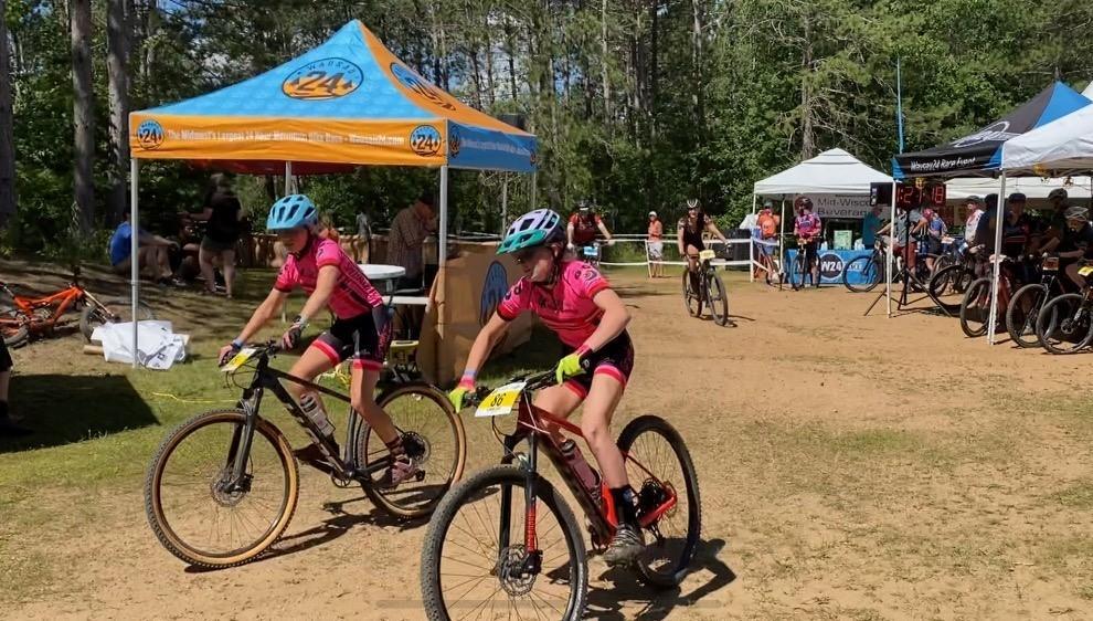 Cyclists in pink jerseys at an outdoor event under blue tents.