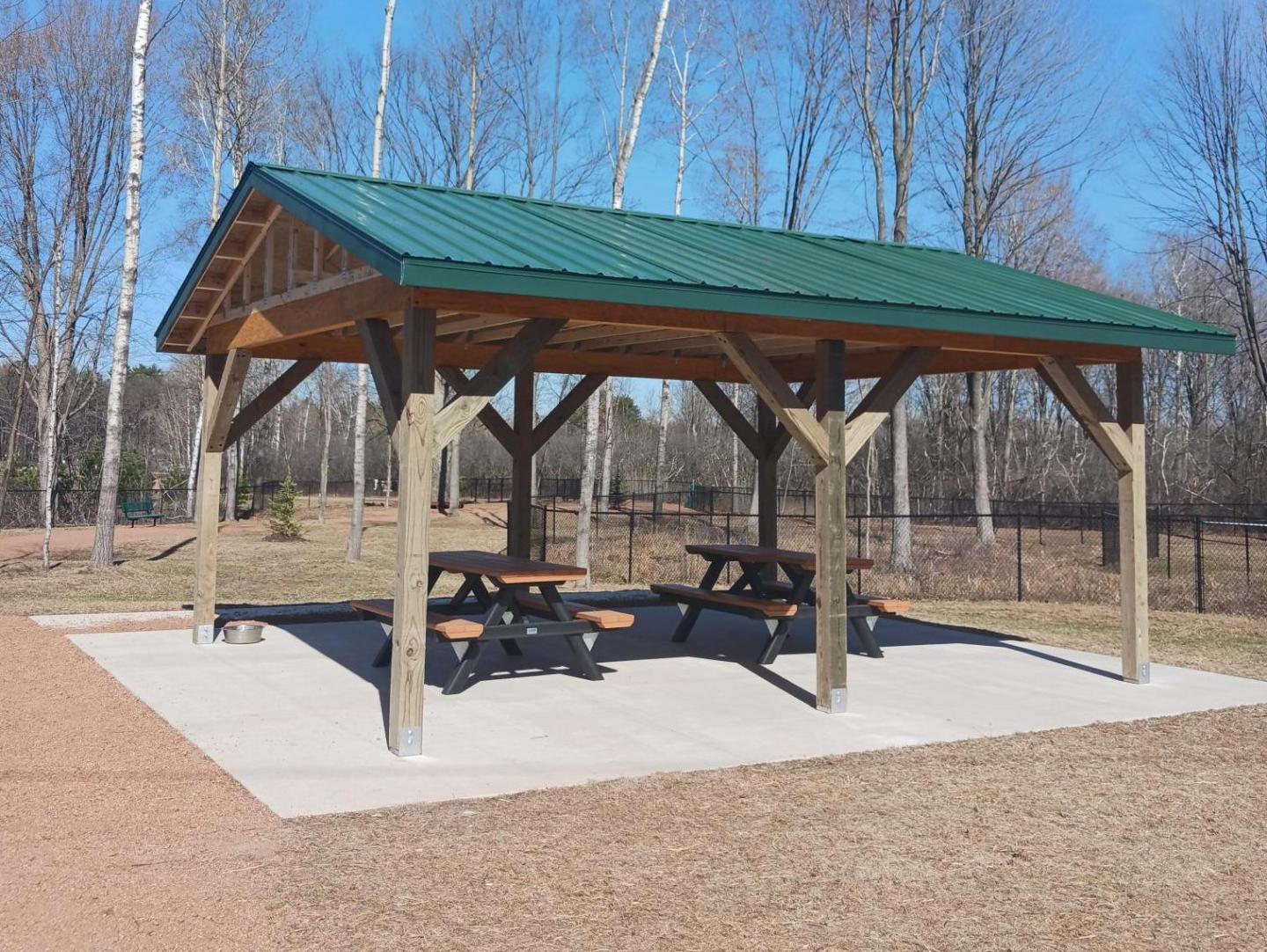 Open wooden shelter with picnic tables under a green roof in a park setting.