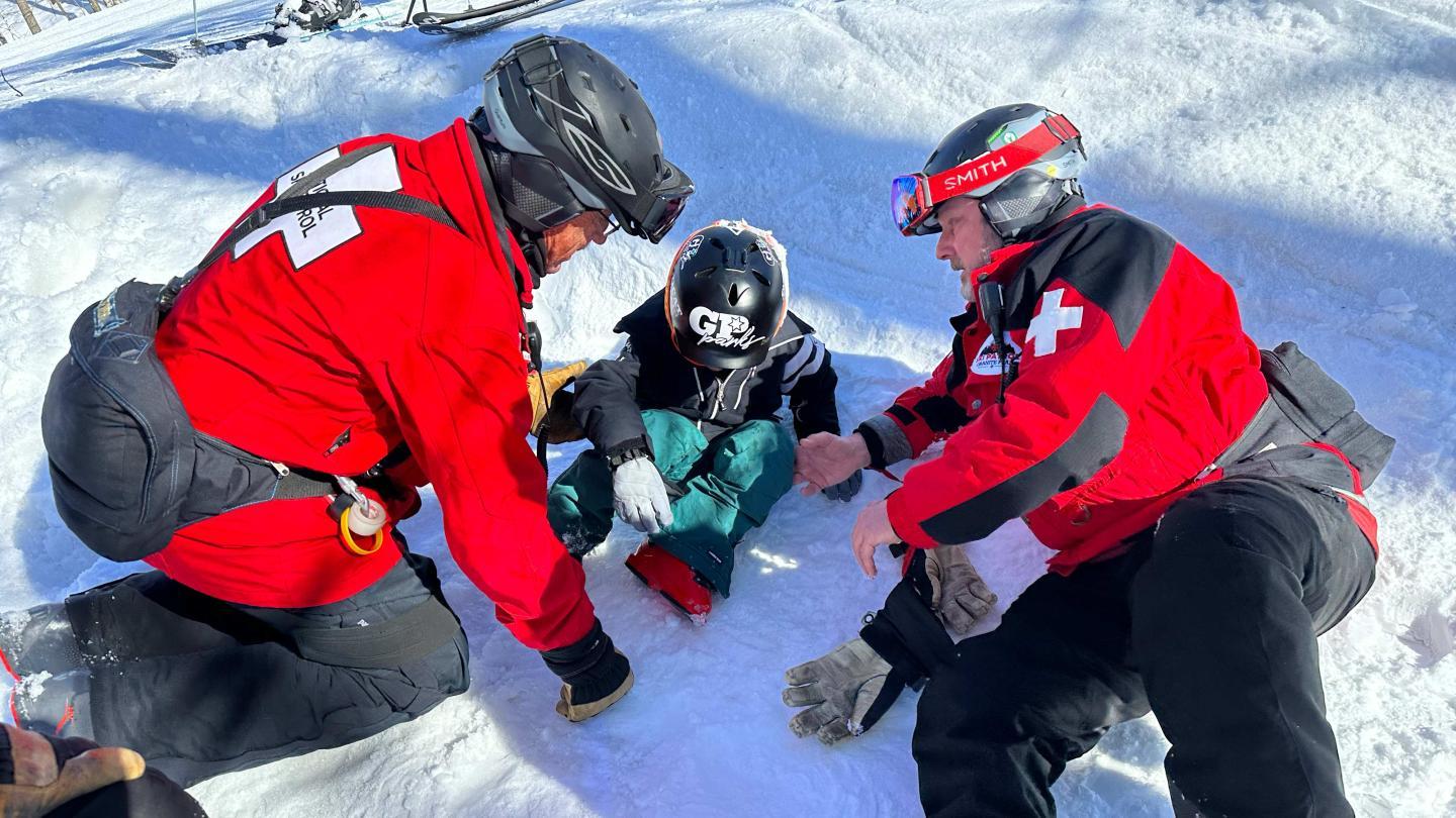 Ski patrol assisting a child sitting in snow, wearing helmets and red jackets.