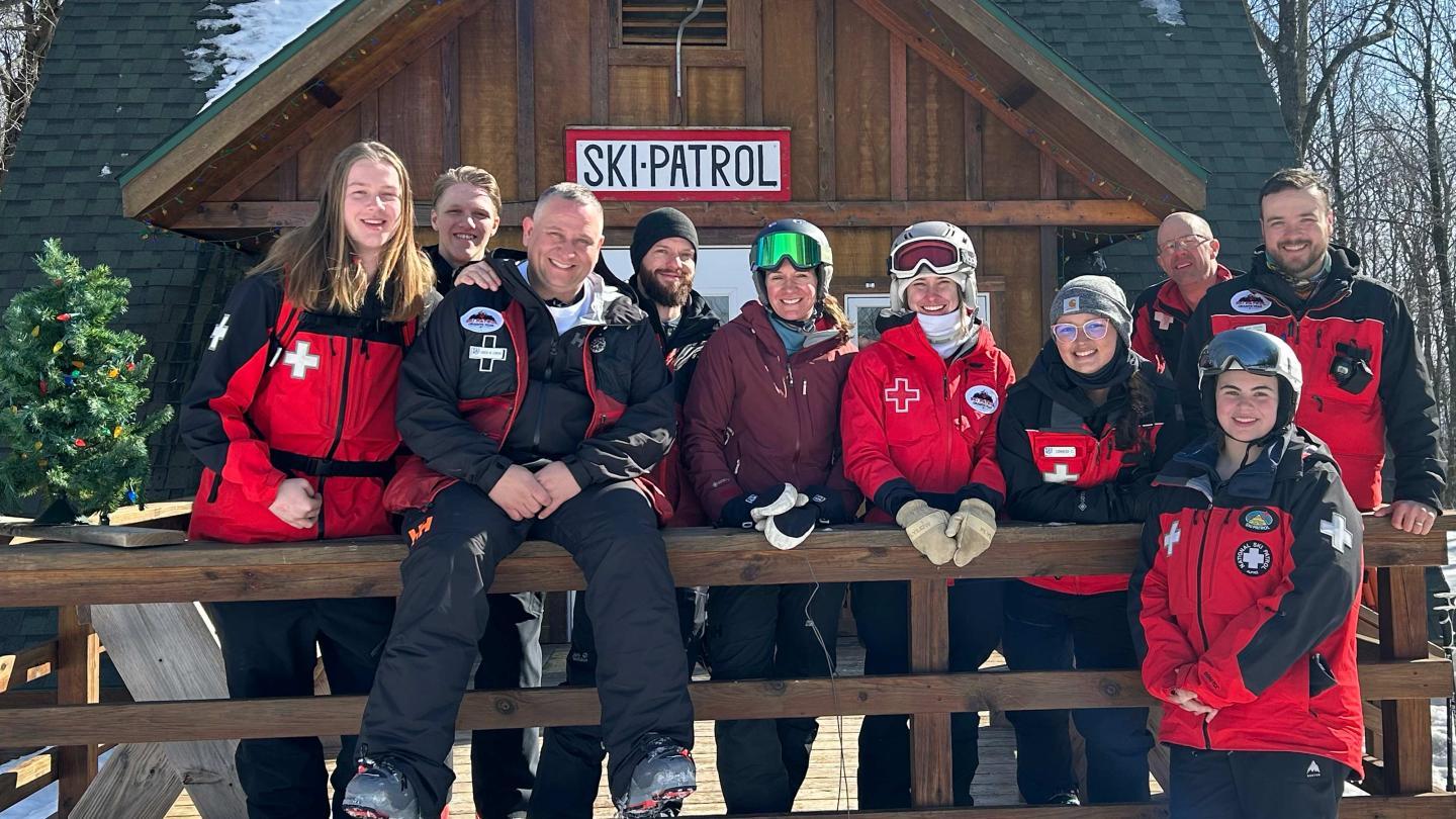 Ski patrol team posing in red jackets outside a wooden cabin in snow.