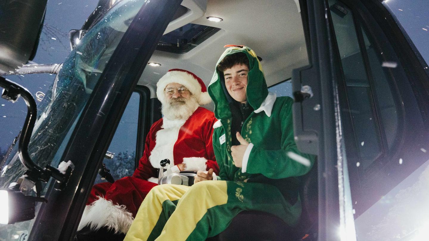Santa and elf sitting in a snow-covered vehicle cabin, smiling.