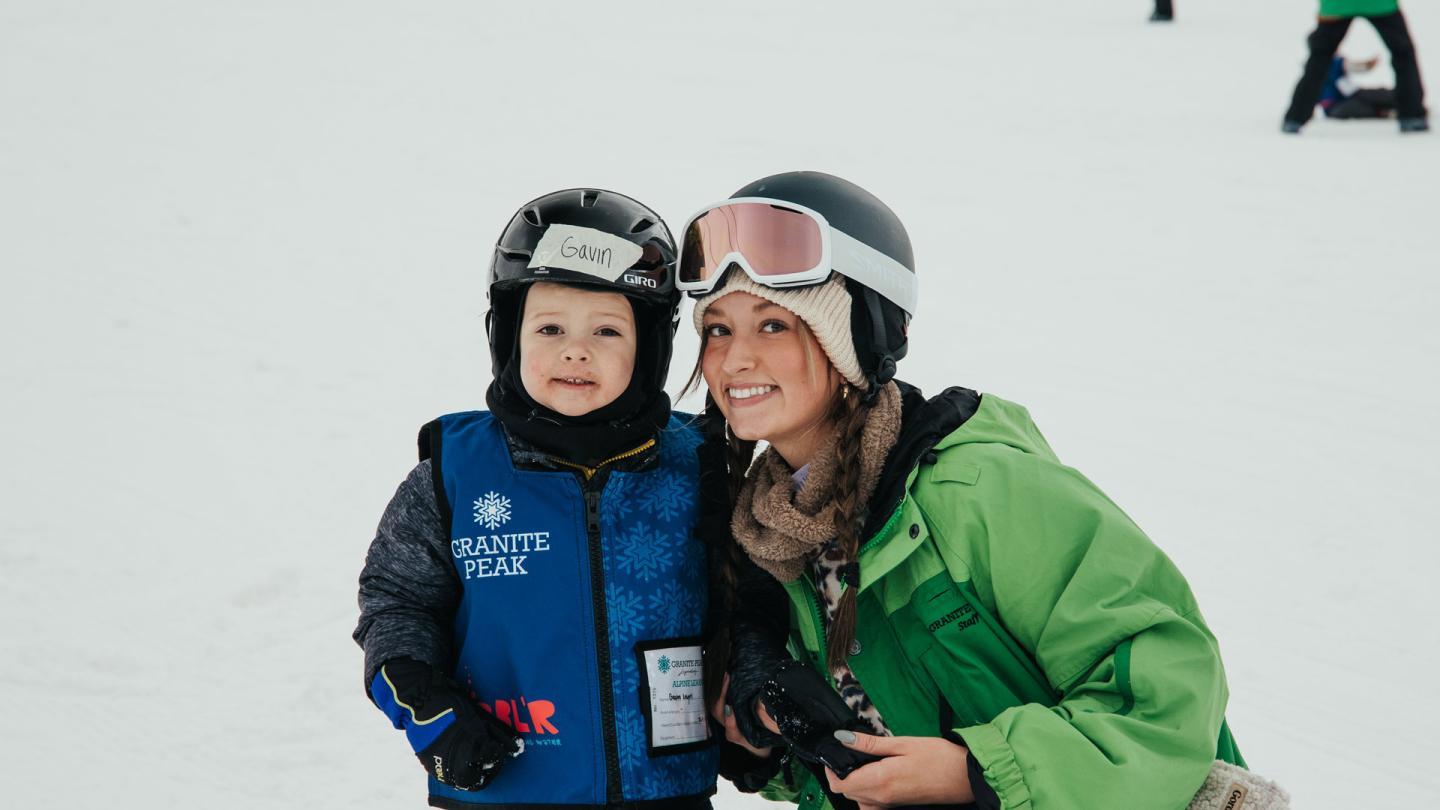 Young child and adult in ski gear smiling on a snowy slope.