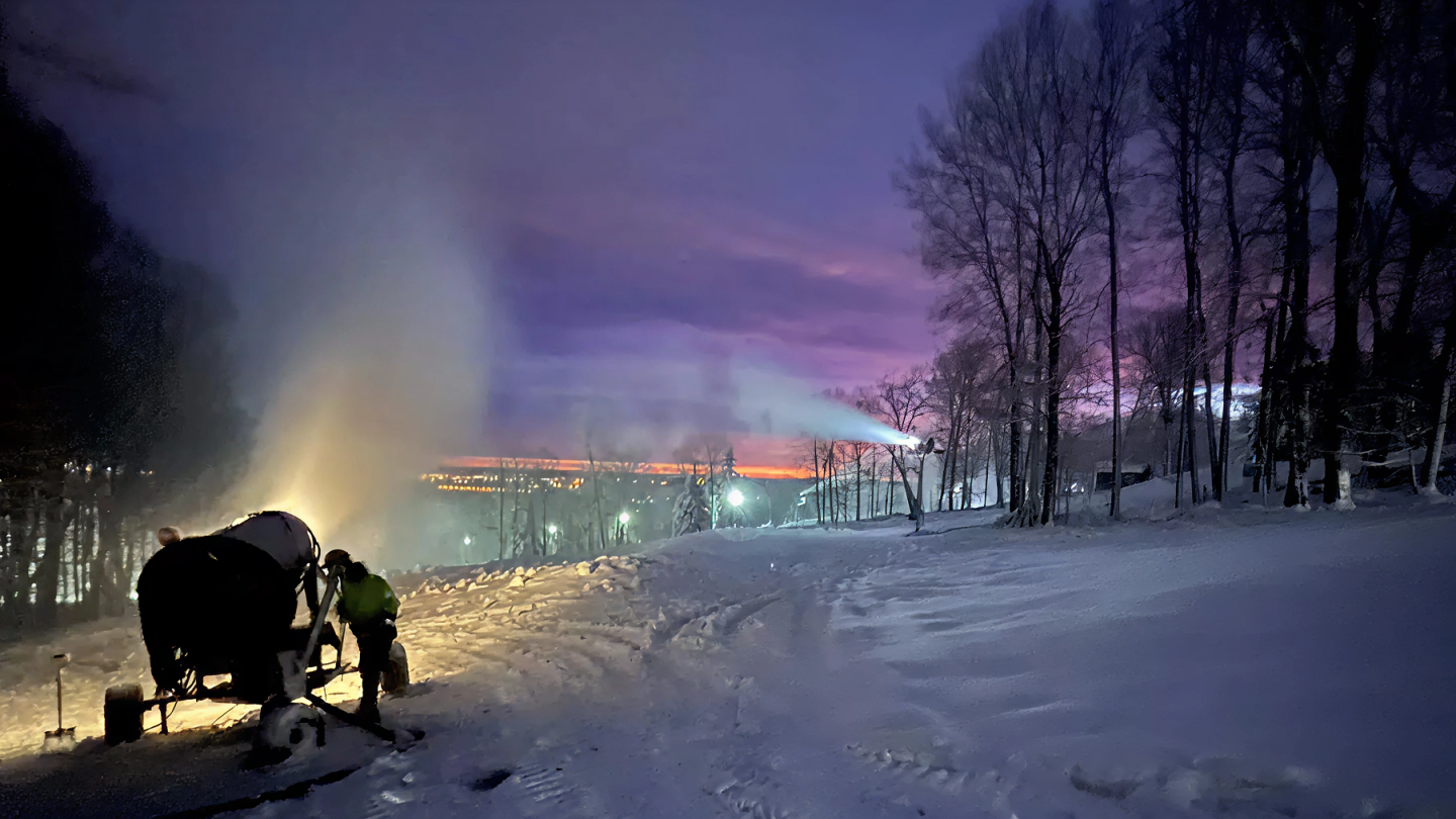 Snow cannon operates at dusk on a snowy slope, with purple sky and silhouetted trees.