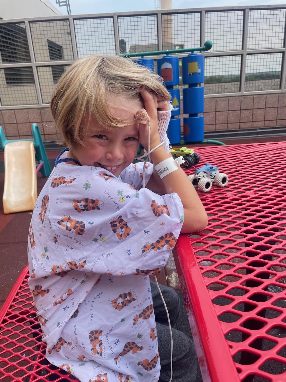 Child in hospital gown sitting at red picnic table, outdoor play area background.