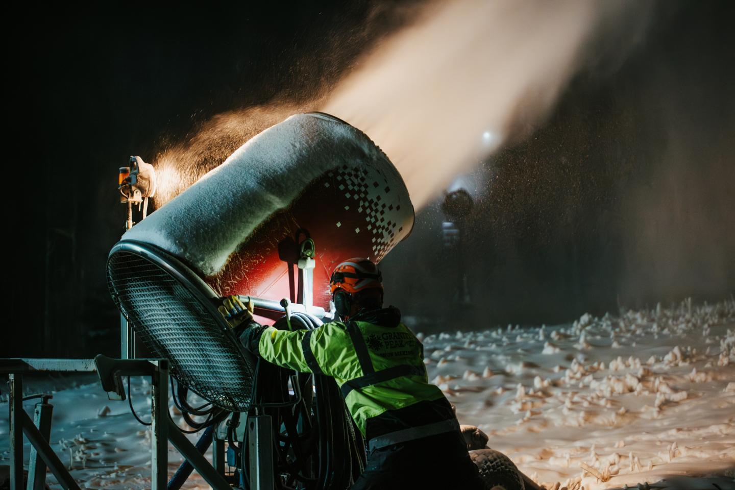 Worker adjusts large snowmaking machine in dim light.
