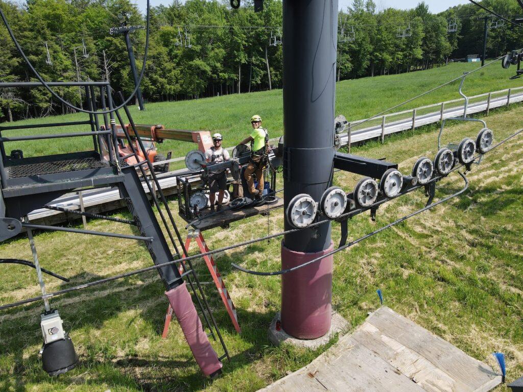 Two workers on a ski lift maintenance platform surrounded by green grass.
