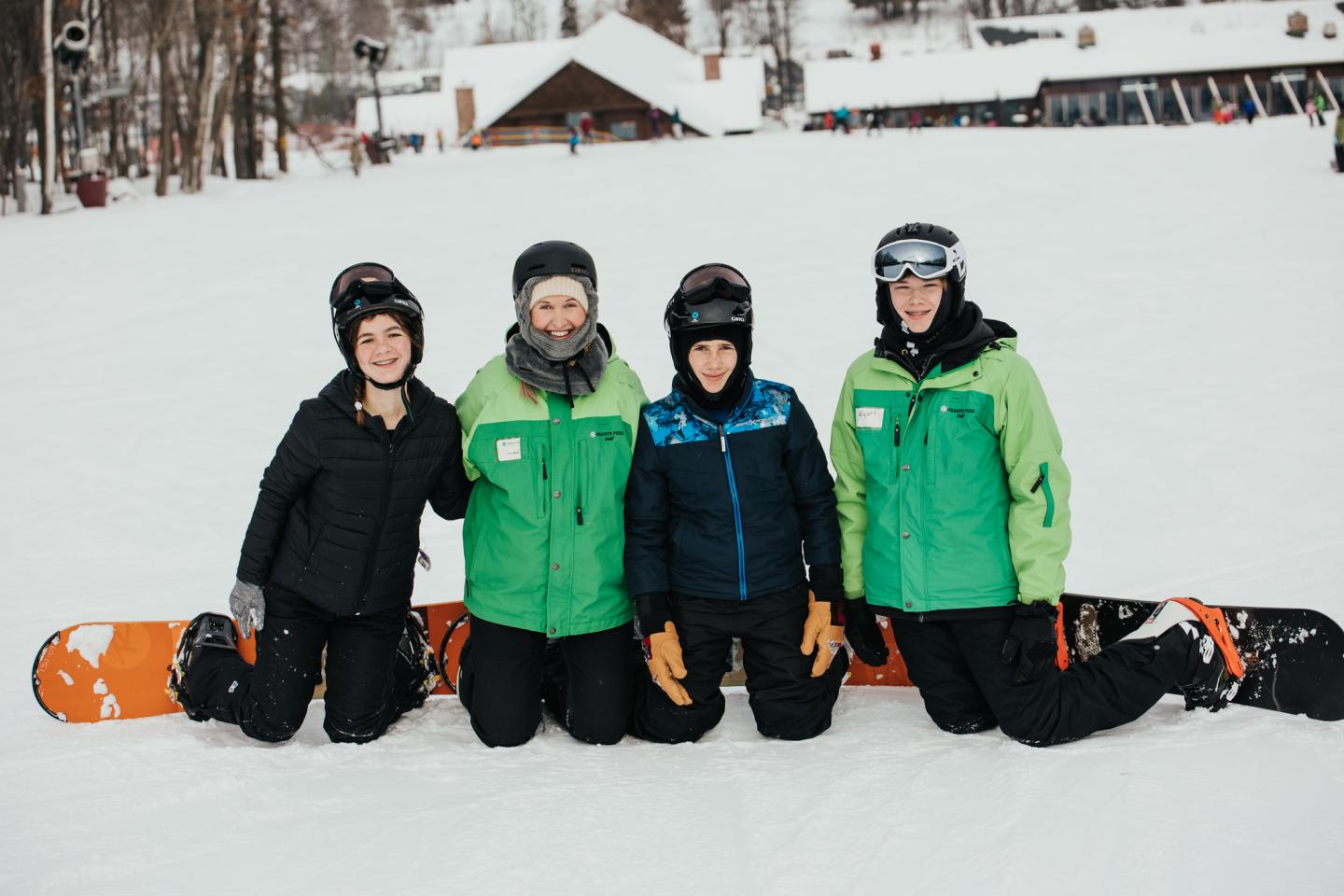 Four people in winter gear kneeling on snow with snowboards, smiling at a ski resort.