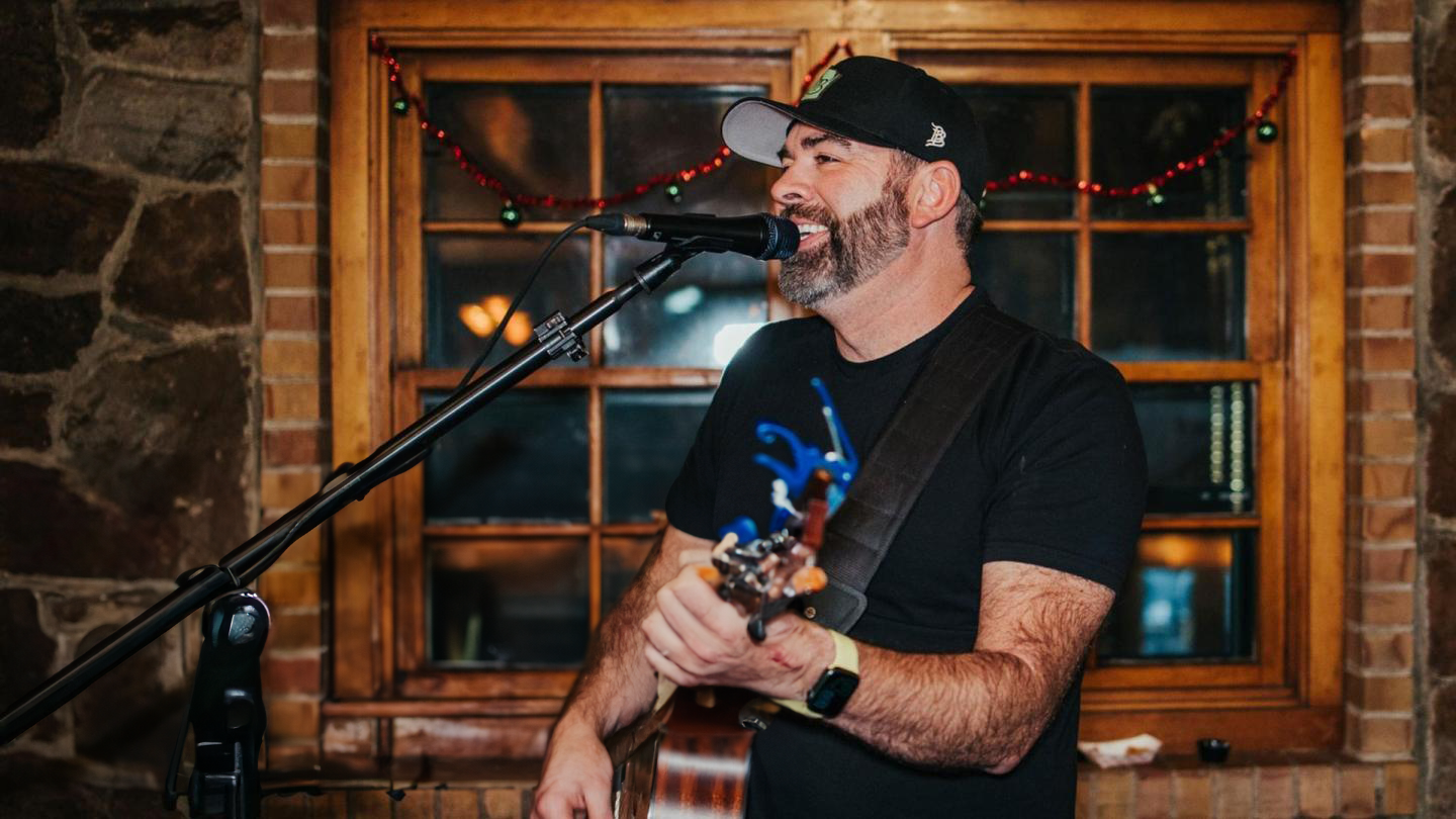 Musician singing and playing guitar indoors, wearing a cap and black shirt.