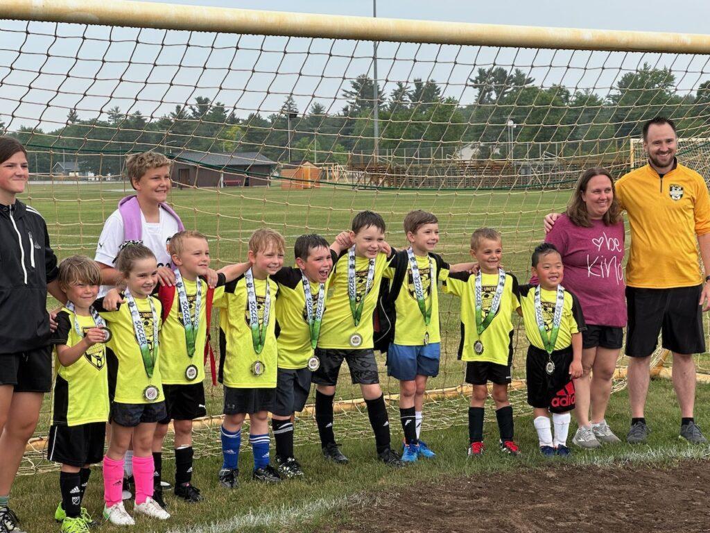 Youth soccer team posing in front of goal, wearing yellow jerseys, with coaches.