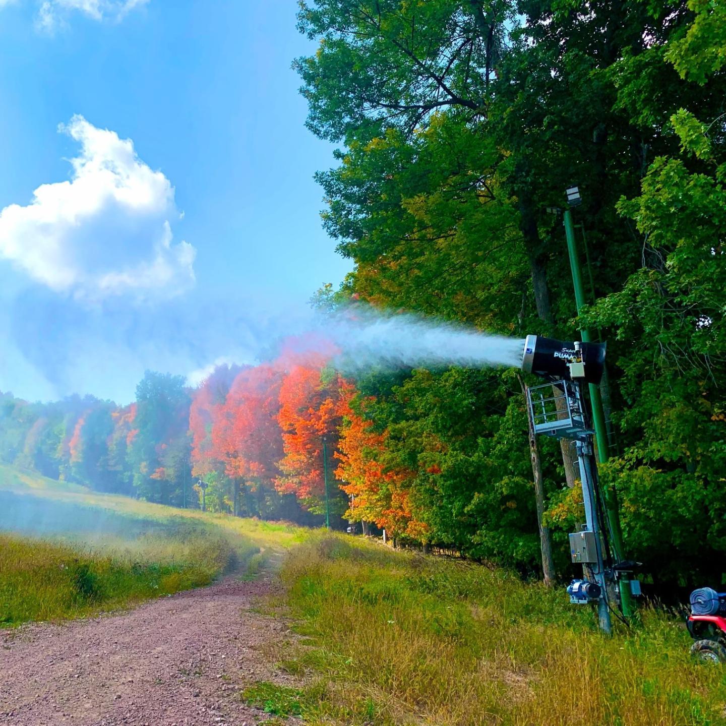 Snow machine in a grassy field with colorful autumn trees and blue sky.