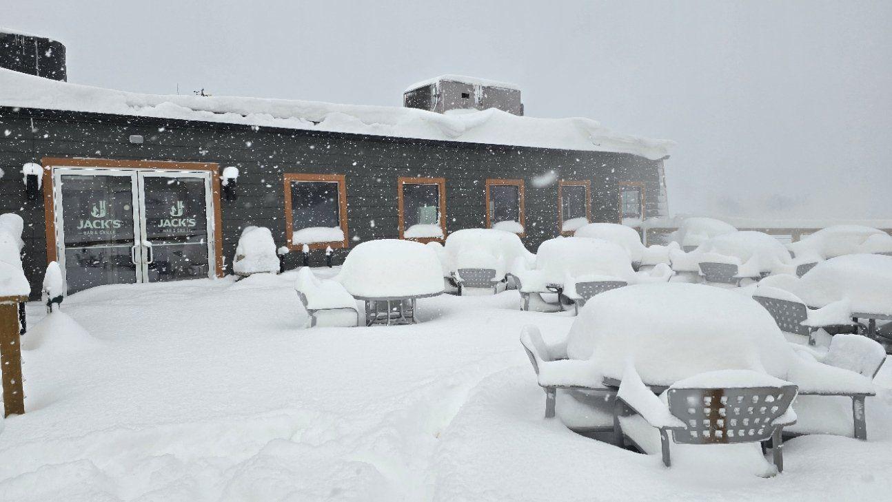 Snow-covered patio with tables and chairs outside a building. Thick snowfall ongoing.