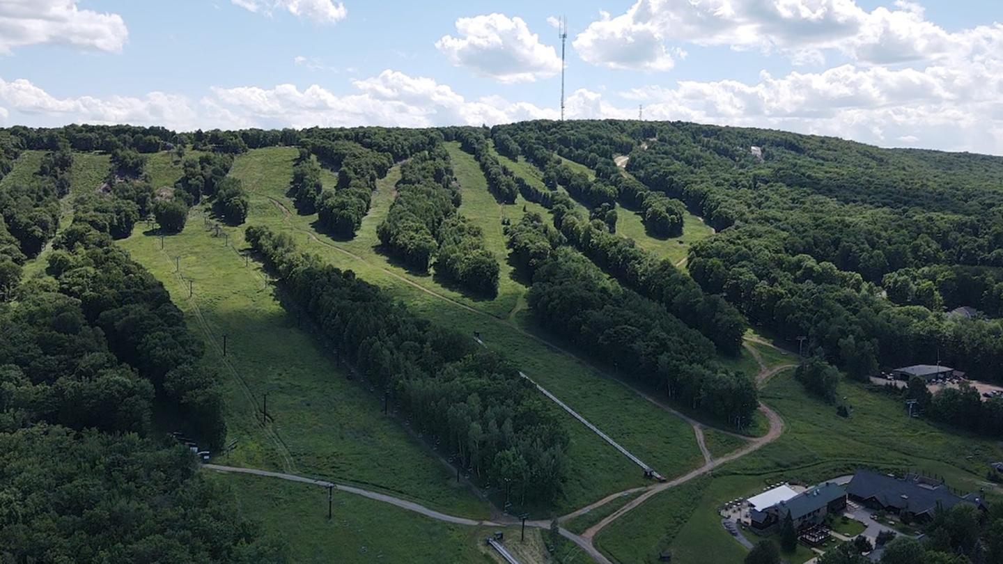 Aerial view of a green hillside with winding trails and scattered trees under a partly cloudy sky.