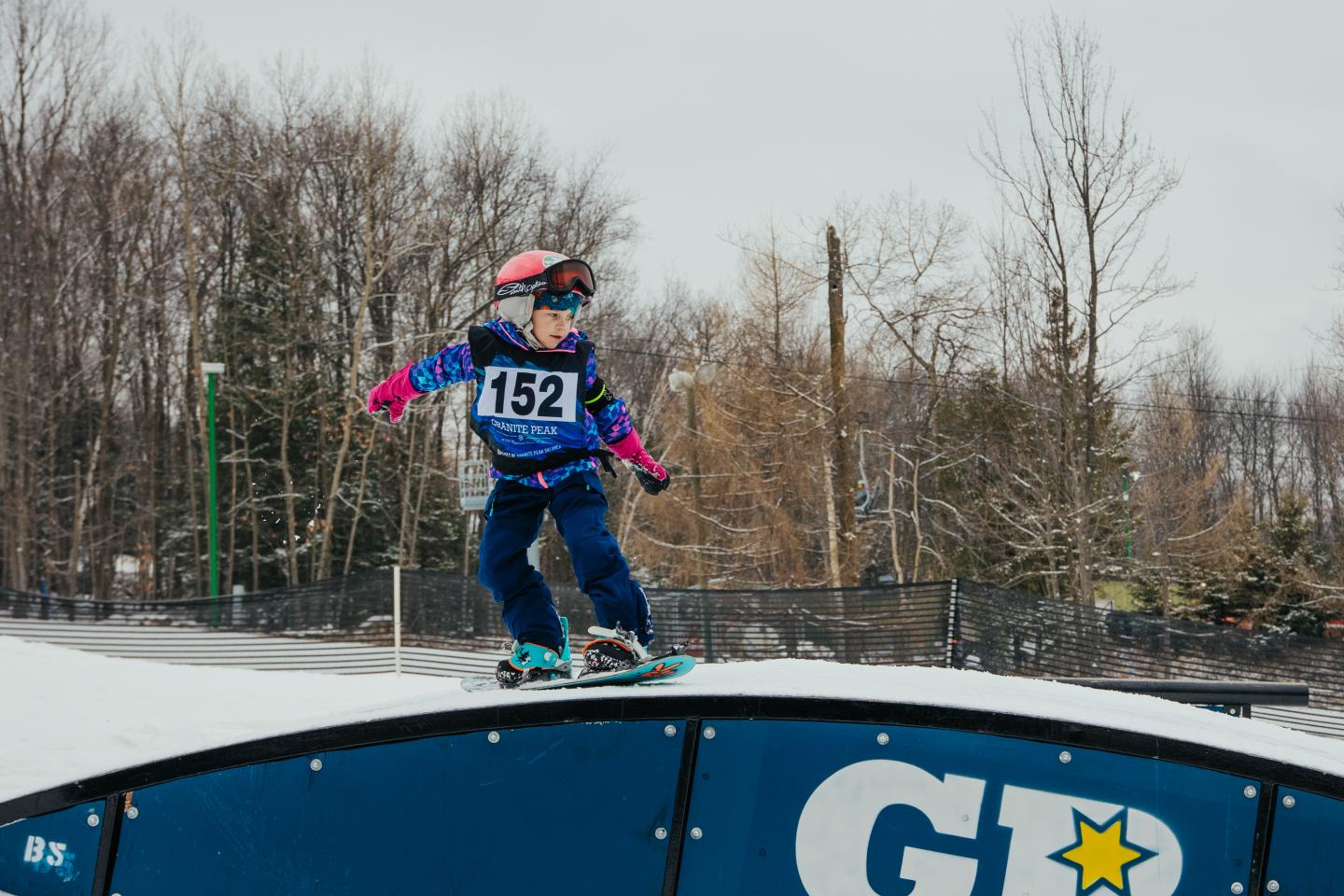 Child snowboarding on a rail, wearing goggles and helmet, overcast day.