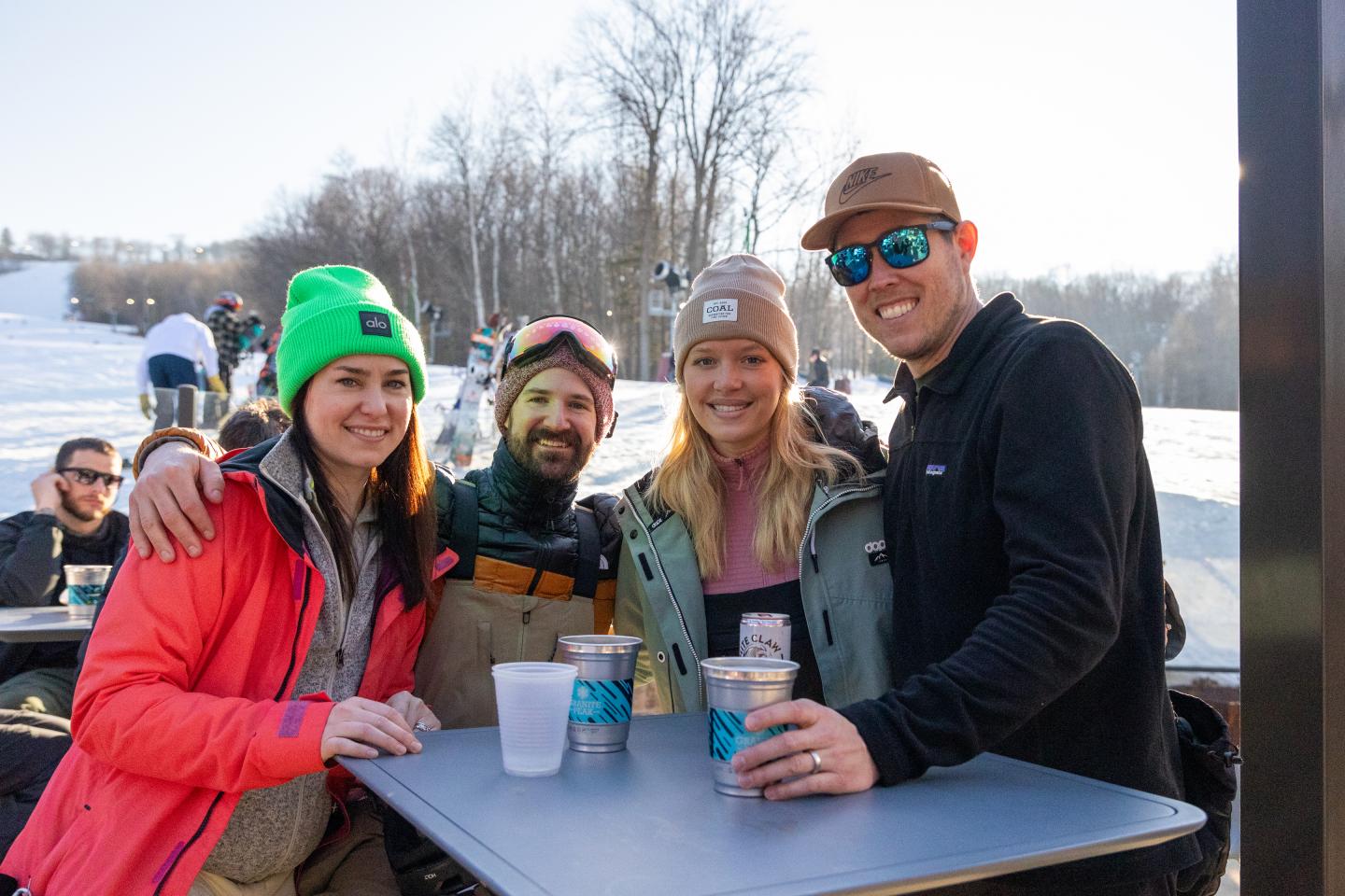 Skiers smiling around an outdoor table with drinks on a sunny, snowy day.