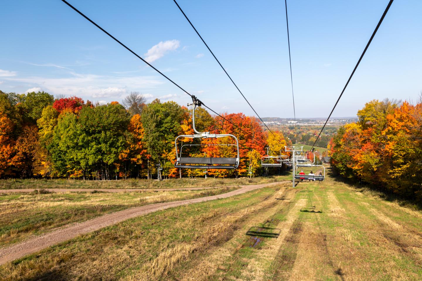 Chairs on a ski lift above grassy slope, surrounded by autumn trees.