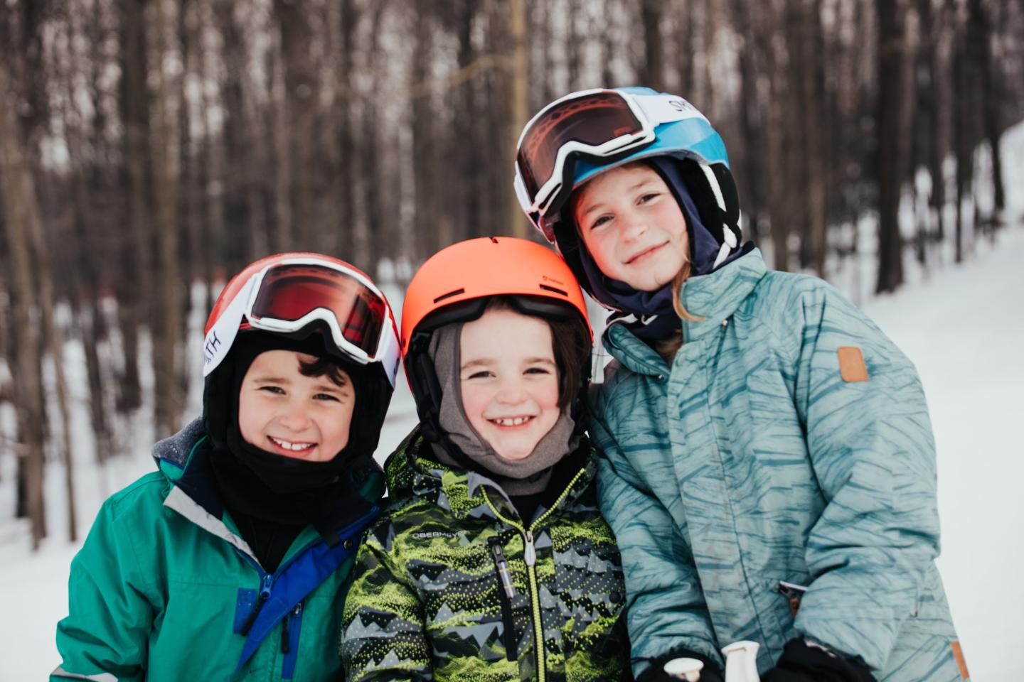 Three smiling kids wearing ski gear and helmets in a snowy forest.