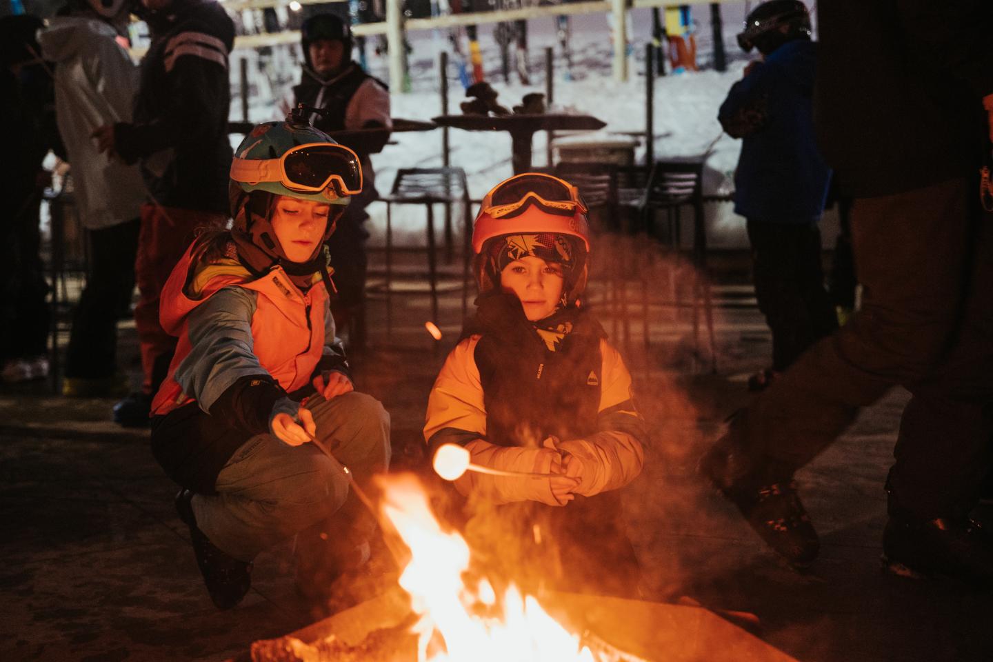Children in ski gear roasting marshmallows over a campfire at a snowy resort.