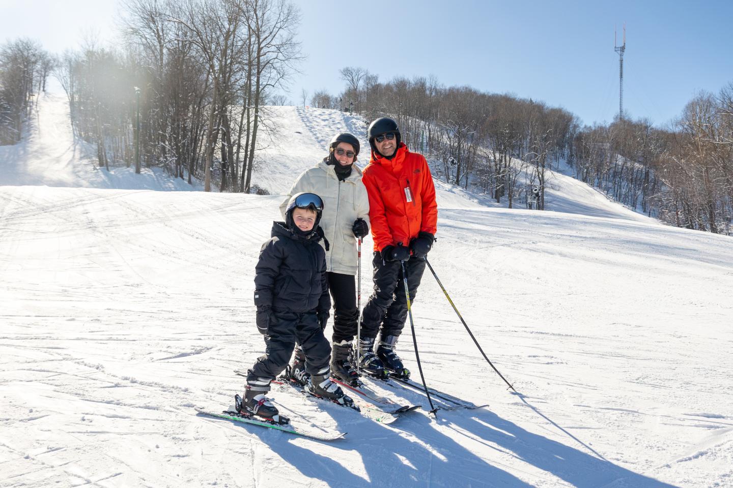 Family skiing on a sunny, snowy slope with bare trees in the background.