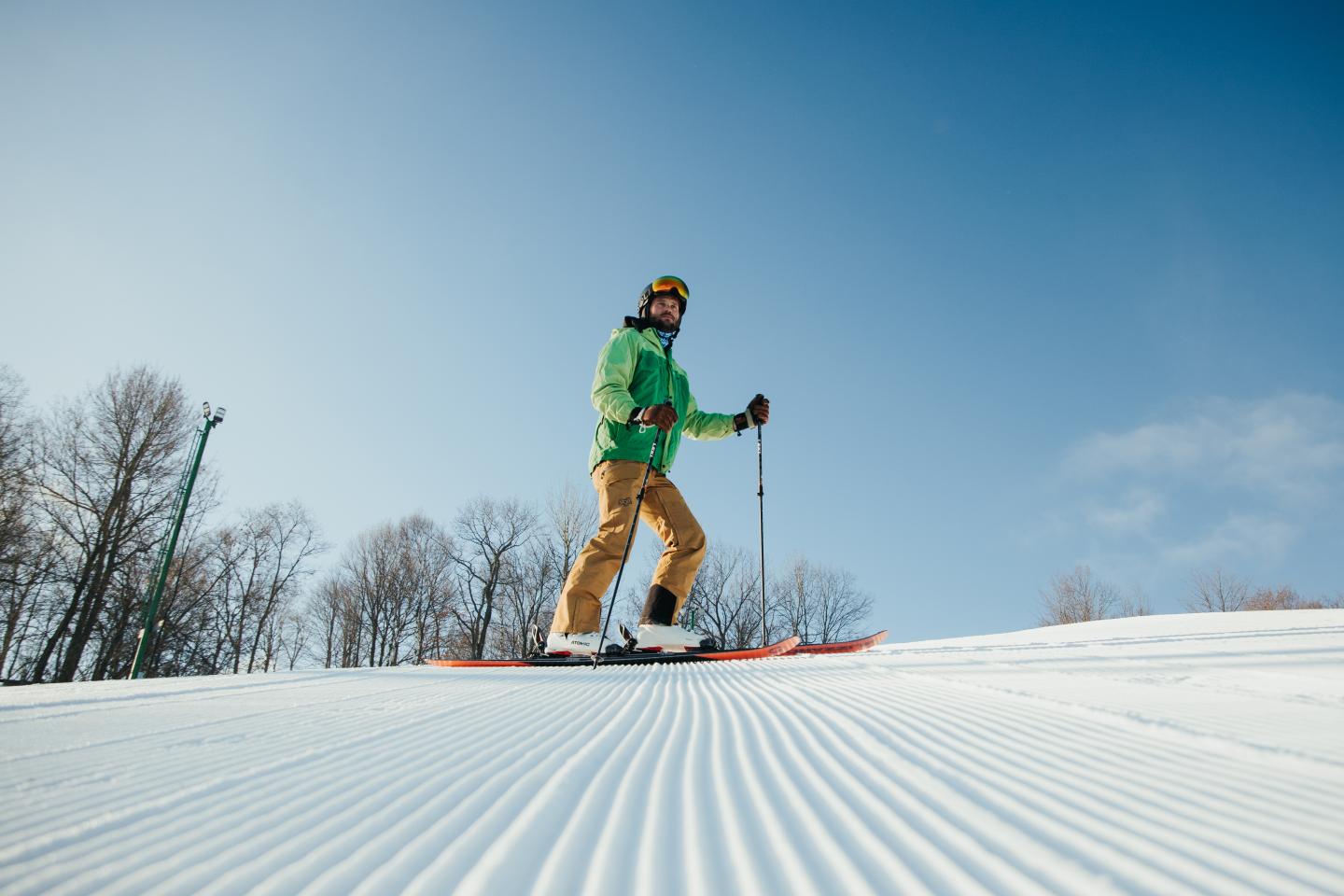 Skier in green jacket on snowy slope under clear blue sky.