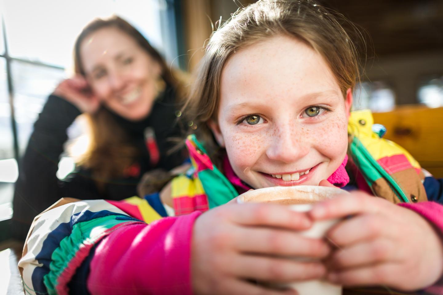 Smiling girl in colorful jacket holding a mug, with a woman smiling in the background.