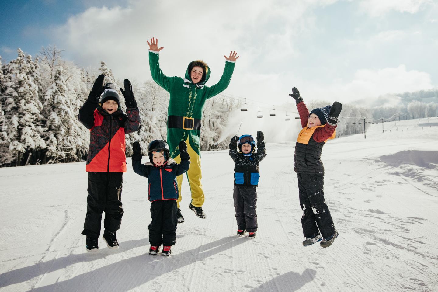 Four children in colorful winter clothes jumping and raising their arms in snowy sunlight.