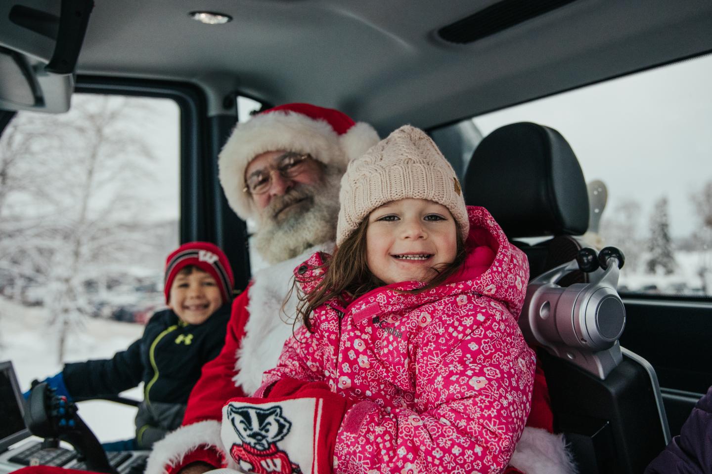 Girl in pink snowsuit smiles beside Santa in a snowy vehicle.